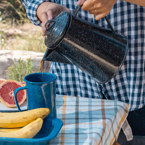 Pouring coffee from 3 Qt Coffee Boiler In Speckled Black For Quick Brews into a blue cup, on checkered cloth with fruits nearby.