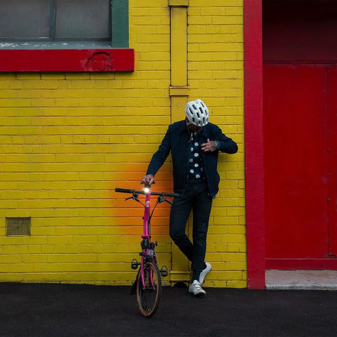 Person leaning on a yellow brick wall with a bicycle equipped with a USB Rechargeable Bike Light With 250 Lumen Front Headlight Ride Ready Low Battery Indicator Side Visibility Cutouts IP67 Waterproof Five Modes And Up To 240 Hours Of Runtime visible on the handlebars.