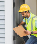 Person in yellow hard hat checks door hinge installation while kneeling with clipboard by the door.