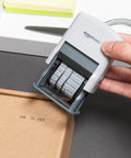 Heavy Duty Self Inking Date Stamp For Office Documents Legal Papers And Corporate Mail Featuring Bold Saturation Marks and a Flexible Ten Year Setting held over a package with date mark.
