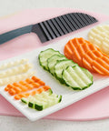 Large crinkle cutter knife with gray handle and black striped blade, beside sliced cucumbers and carrots on a plate.