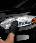CERAKOTE Ceramic Headlight Restoration Kit being applied to a car headlight by gloved hand on a black vehicle.