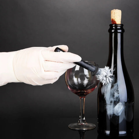 A person wearing disposable latex exam gloves cleaning a wine bottle with a brush and a glass of red wine nearby.