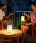 Two people on patio with Cordless LED Lantern Table Lamp With Remote, IP44 Shower Safe And Waterproof, providing warm light.