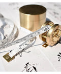 Top Shelf Vintage Brass Mini Tape Measure With Retractable Sewing Ruler That Is Totally Built Tough For All Tailor Jobs Featuring One Meter Gauges on a table with printed designs.