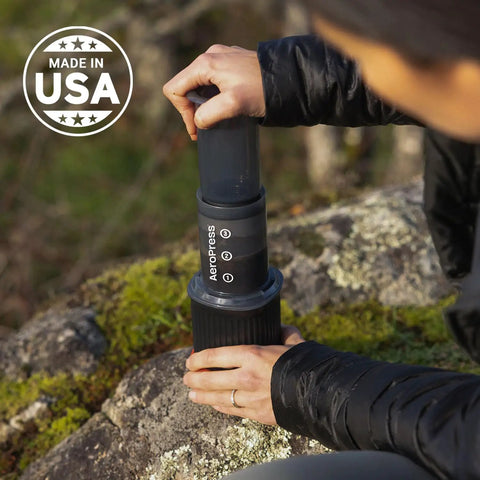 Person using Ultra Portable Travel Coffee Maker Kit For Grab And Go Outdoors All In One French Press Pour Over And Espresso Style Manual Brewer For Camping And Hiking Two Minute Brew For A Less Bitter More Flavorful Cup on a rock.