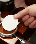 Top Shelf Espresso Barista Gear Featuring 54 Mm Stainless Steel Tamper Fits Breville Portafilters Includes Precision Wdt Needles Sleek Dosing Funnel And Two Hundred Premium Paper Filters To Nail That Perfect Morning Pull, showing a hand placing a paper filter on portafilter.