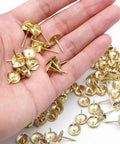 Slick Daisies Antique Gold Upholstery Tacks And 100 Piece Bronze Flat Head Nails With Rust Proof Finish in a hand, showcasing the antique gold color and design.