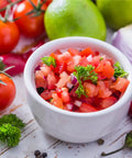 Bowl of fresh salsa surrounded by tomatoes, limes, chili peppers, cilantro, and onions on a rustic white surface.