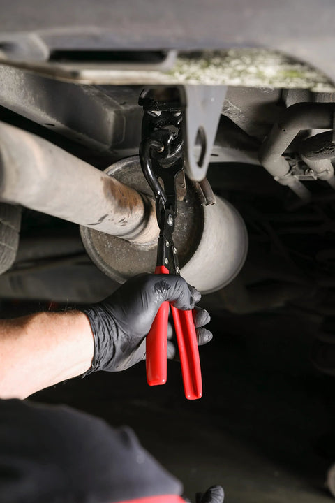 Under-vehicle view of ARES 14020 heavy-duty exhaust hanger removal pliers with red handles gripping a muffler hanger