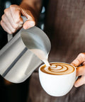 Four Pack 20 Oz Stainless Steel Milk Frothing Pitchers For Coffee And Latte Art Barista Gear Steamer Jugs Cappuccino Maker Pro Grade Stainless Steel Home Cafe And Shop Essentials being used to pour milk into a cappuccino cup with latte art.