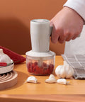 Cordless Electric Hand Mixer With Stainless Steel Beaters. USB Rechargeable Power For Quick Whipping And Blending Eggs Or Cream. Three Speed Settings Ensure Smooth Mixing Every Time. Ergonomic Grip Makes Baking Easy And Fun For All Skill Levels At Home. Hand using a cordless mixer on garlic and peppers.