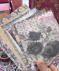 Hand holding floral scrapbook paper from All In One Vintage Junk Journal Kit With 185 Pieces Scrapbook Paper Pack A6 Aesthetic Notebook Scrapbook Kit Art Craft Diy Gift For Adults Women Starter Friendly in front of embellishments and patterned book cover in background.