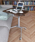 Totally Adjustable Pneumatic Sit Stand Rolling Laptop Desk Cart with walnut surface and silver alloy base next to a couch.