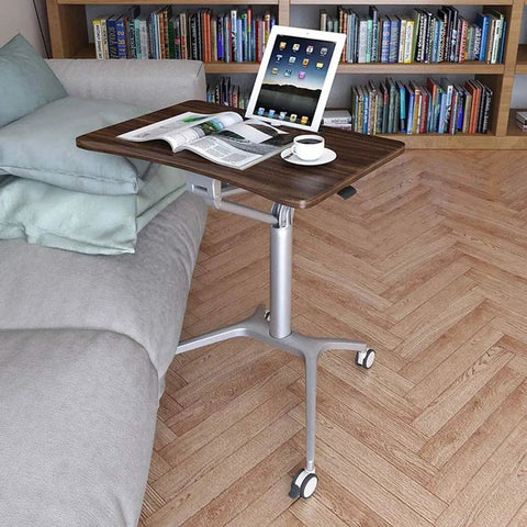 Totally Adjustable Pneumatic Sit Stand Rolling Laptop Desk Cart with walnut surface and silver alloy base next to a couch.
