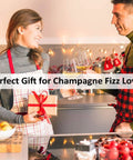 Two people toasting with champagne in a decorated kitchen, wearing aprons, holding a gift, promoting 2 Pack Champagne Stoppers With Built In Vacuum Pump Black Keeps Bubbles Fresh To The Last Drop And Keeps It Fizzy With A Game Changing Pump Piston System For A Tight Seal And Leak Proof Sparkling Wine Saver.