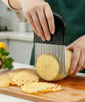 Black-handle potato cutter with stainless steel wavy blade slicing a potato into ridged fries.