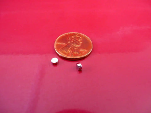 Tiny 3x1.5mm Neodymium Disc Magnets Pack Of 300 For Warhammer And Miniatures next to a penny on red background.