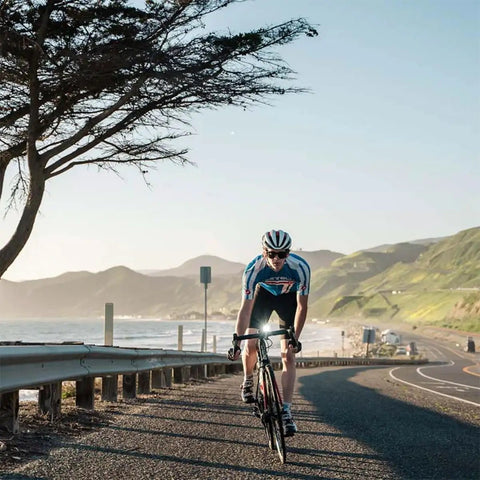 Cyclist on coastal road with Lumina 1000 Boost Front Bike Light With Sabre 110 Rear Light, mountain backdrop