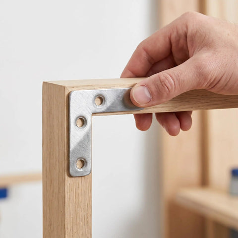 Hand holding 100-Piece Stainless Steel L-Shape Corner Bracket Set for 2x2 Inch Small Angle Joints on wooden frame