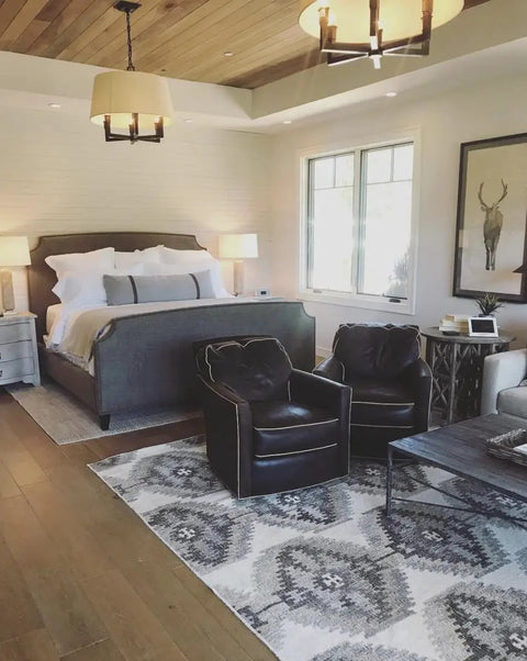 Cozy bedroom featuring a gray upholstered bed, leather chairs, and a decorative rug, showcasing warm indoor aesthetics.