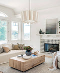 Modern living room with white pendant light, beige sofa, wood coffee table, and patterned fireplace. Large windows and decor accents.