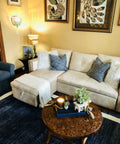 Cozy living room with beige sectional sofa, decorative cushions, round wooden coffee table, wall art, and vintage wall lamp.