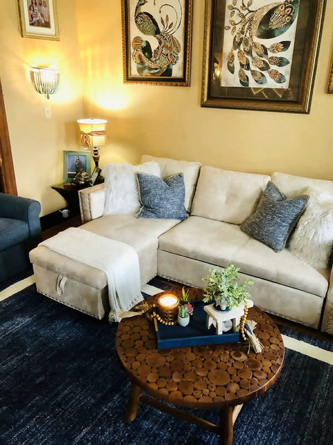 Cozy living room with beige sectional sofa, decorative cushions, round wooden coffee table, wall art, and vintage wall lamp.