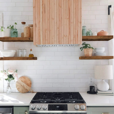 Kitchen with wooden range hood and floating shelves with decor items on white tile backsplash