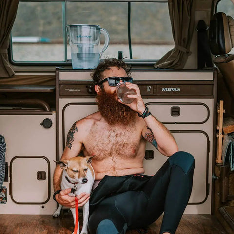 Man sitting in camper van with dog, drinking water, Pure Filter Water Pitcher For Refrigerator 10 Cup Capacity visible on counter.