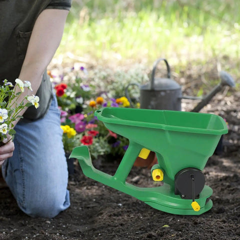 Person gardening with the Effortless Hand Crank Seed Spreader 1.5L for Garden and Lawn placed on soil, surrounded by flowers.