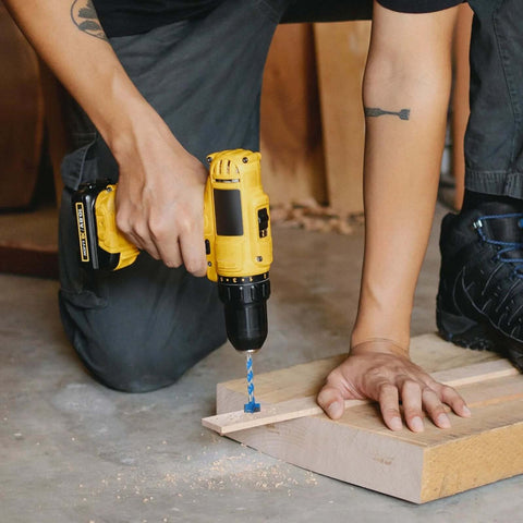 Side view of a yellow cordless drill with a blue twist drill bit drilling into a wooden block