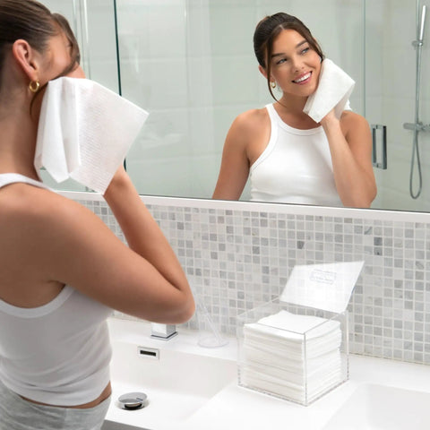 Woman using a facial wipe in front of a mirror with the No Fuss Clear Acrylic Storage Box For Towels And Towelettes Countertop Dispenser Bin.