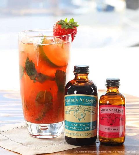 Glass of iced beverage with cucumber and strawberry garnish next to Nielsen-Massey Tahitian vanilla and rose water bottles.