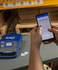 Hella Tough Portable Label Printer With Auto Cut And Badass Build with smartphone in use, printing fast text and barcodes in a warehouse setting.