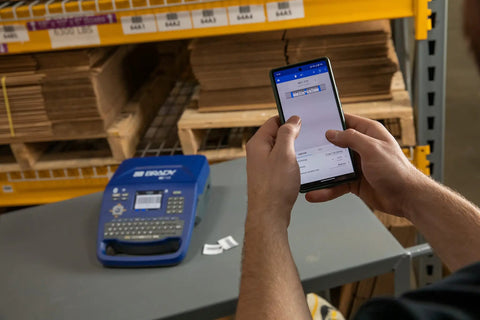 Hella Tough Portable Label Printer With Auto Cut And Badass Build with smartphone in use, printing fast text and barcodes in a warehouse setting.