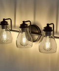 Black Three Light Vanity Light Fixture With Clear Glass Shades For Bathroom Over Mirror, warm glow on wall