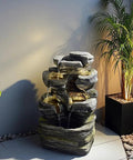 Six Tier Cascading Rock Outdoor Water Fountain With Lights For Garden Patio Backyard Deck And Porch No Fuss Setup With Pump 24.4 Inch High 110V Plug, shown installed with decorative plants.