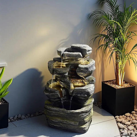 Six Tier Cascading Rock Outdoor Water Fountain With Lights For Garden Patio Backyard Deck And Porch No Fuss Setup With Pump 24.4 Inch High 110V Plug, shown installed with decorative plants.