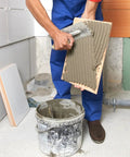 Person applying adhesive to tile, bucket of grout nearby, in bathroom renovation setting.