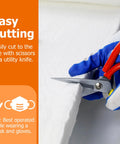 Close-up of gloved hands cutting a white ceramic fiber insulation blanket with red-handled scissors.