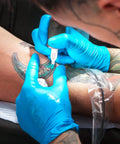 Tattoo artist using a tattoo machine on a client's arm wearing Heavy Duty Disposable Nitrile Gloves Large Size 4 Mil Thick.