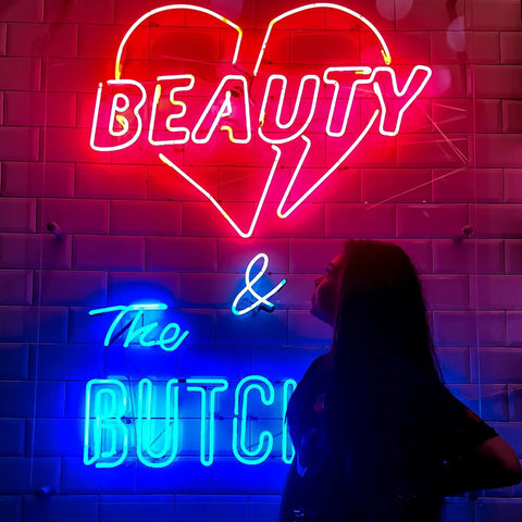 Woman standing in front of colorful neon sign reading 'Beauty & The Butch' with heart shape, showcasing vibrant lighting.