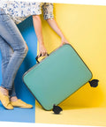 Person carrying a green suitcase with black wheels on blue and yellow background, showcasing stylish travel gear.