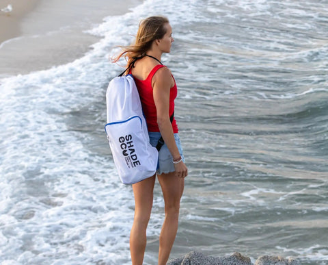 Person carrying Easy Setup Beach Canopy Tent 8.5x9 Ft UPF50 Plus Sun Shade Portable Four Post Shelter on a beach in white carry bag.