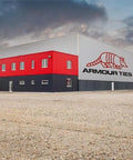 Exterior view of a large red and gray industrial building with 'Armour Ties' logo, situated in a gravel lot under a cloudy sky.