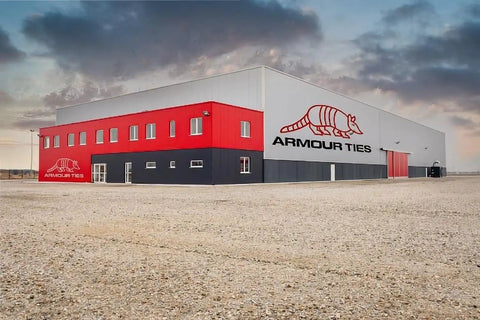 Exterior view of a large red and gray industrial building with 'Armour Ties' logo, situated in a gravel lot under a cloudy sky.
