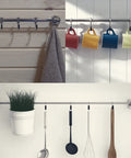 Metal rod with hanging hooks holding towels, cups, utensils, and a plant pot on a light wooden and white ceramic background