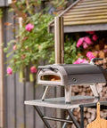 Outdoor pizza oven setup on a table with wood logs and flowers in the background.
