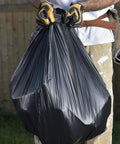 Person holding a large Built Tough Black Trash Bag 55 To 60 Gallon 1 Point 5 Mil Thick ready for disposal outdoors.